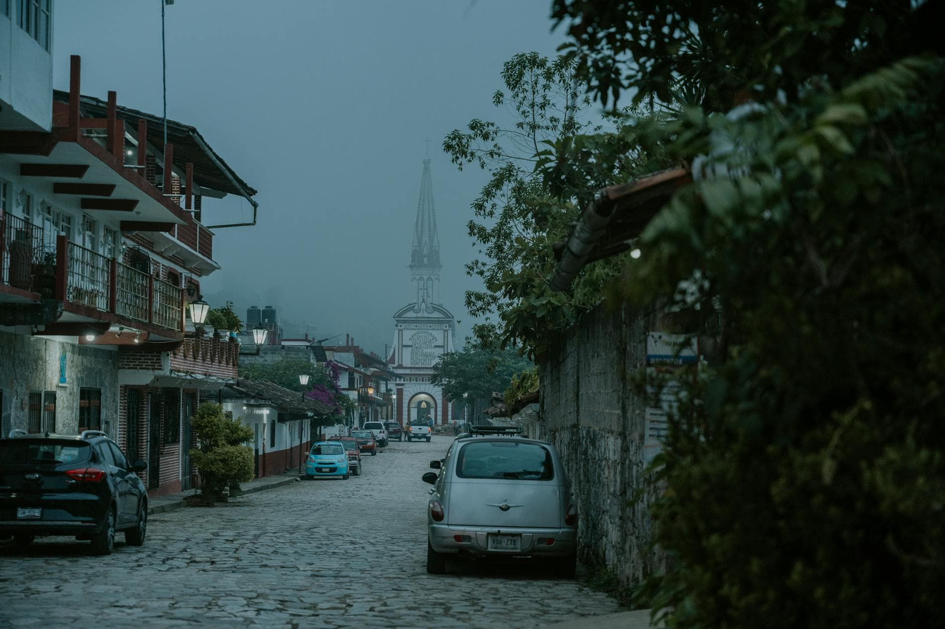 Scenic cobblestone street in Mexico with parked cars and a historic tower obscured by fog.